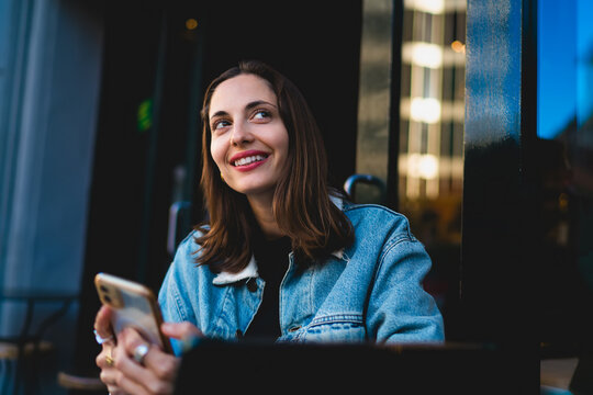 Serene Caucasian female student holding smartphone at outdoor cafe table looking up thoughtfully, listening to audiobooks literature podcast streaming apps during relaxing study break in city