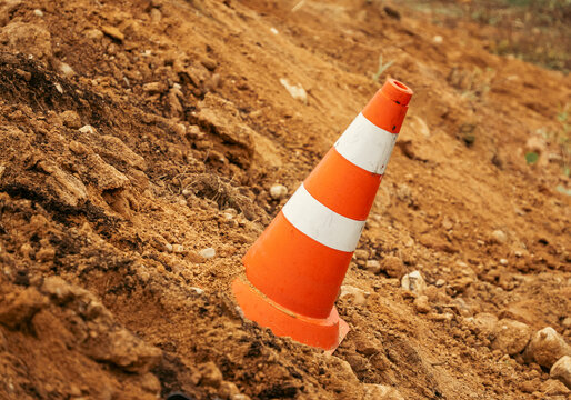 An orange traffic cone with white reflective stripes stands partially embedded on a sandy soil slope at an outdoor construction site.