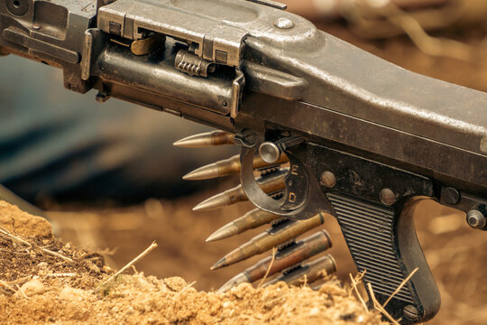 A detailed close view of a historic metal machine gun receiver and pistol grip loaded with a belt of brass cartridges in an outdoor trench.