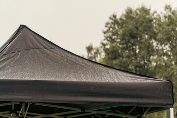 The dark fabric peak of a popup canopy tent is covered in fine raindrops against a blurry background of green trees. © Dmitry