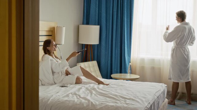 Young adult Caucasian woman switching TV channels in bed and joining man by window in hotel. Suitable for luxury resort advertising and romantic getaway campaigns