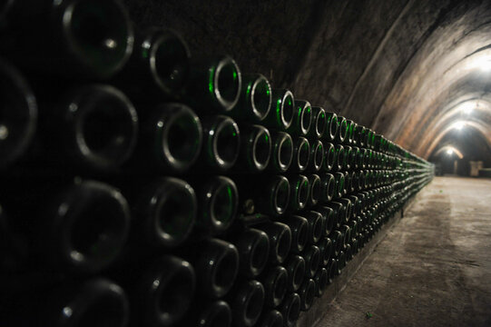 Rows of green glass bottles aging horizontally inside a dark underground wine cellar with arched stone vault ceiling and dim atmospheric lighting along tunnel.
