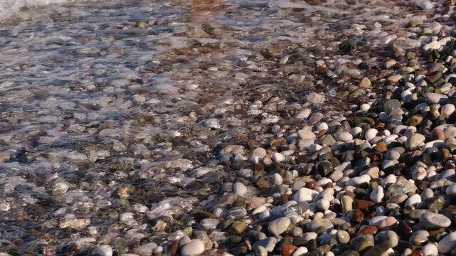 Waves continuously wash over a rocky beach, exposing colorful pebbles and creating foam as the tide ebbs and flows along the shore