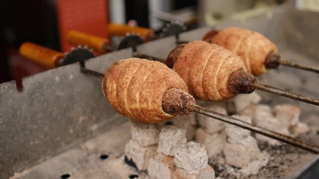 Sweet pastries on skewers are rotating over hot coals in a street food environment, highlighting the cooking process and delicious appearance, trdelnik