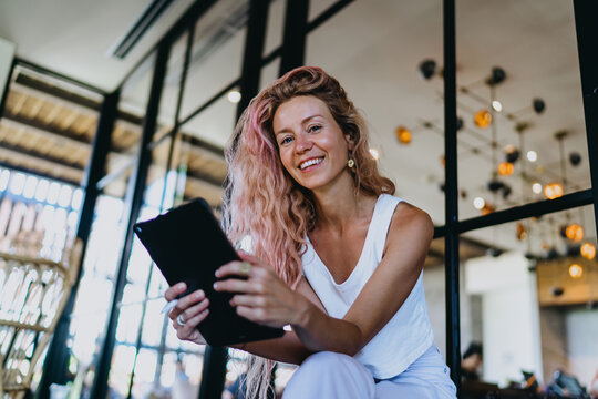Caucasian female web designer holding touch tablet with stylus pen, looking at camera with friendly smile while planning application mockup in modern workspace