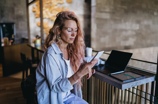 Focused Caucasian female user texting on smartphone while reviewing two factor security verification code, sitting near laptop computer in modern open plan lounge coworking studio