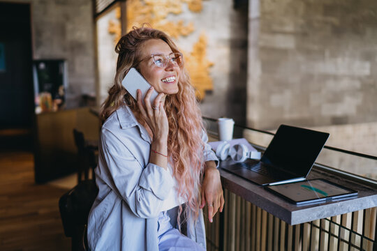 Caucasian female student in glasses laughing during mobile phone call near open laptop and tablet, discussing remote e-learning course assignment in modern lounge workspace studio.