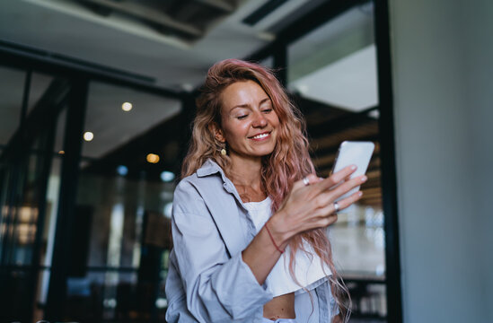 Caucasian female student smiling gently while checking mobile banking application notification on smartphone screen, managing online e-commerce transaction in modern lounge coworking studio