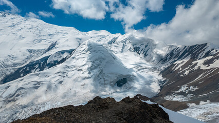 Yukhin Peak stands proudly in Kyrgyzstan with its snow-covered slopes. The view captures the vast mountains and a blue sky. The rugged terrain shows the beauty of nature at this location. © Evgeniy