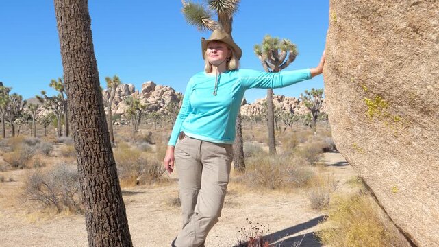 Happy adult woman wearing hat and hiking gear enjoying sunny weather while exploring beautiful and arid landscape of joshua tree national park in california. Woman stands next to a huge boulder.