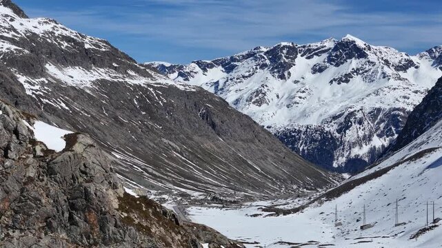 rocky outcrop frames snowbound alpine valley winding road threads floor below scattered pylons trace route power lines cross slopes julier pass mountain crossing graubunden switzerland high altitude