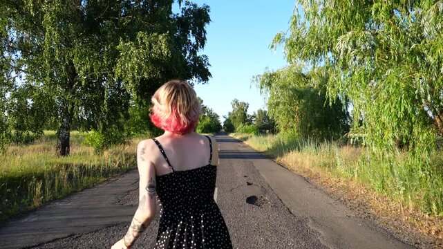 Carefree hippie woman in black dress walking along empty road at countryside. Happy punk girl with pink hair fooling around while goes on country driveway. Young female hipster having fun outdoor