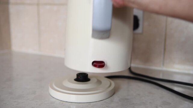 A hand turning on the electric plug switch, putting a kettle on its base and pressing the on button to boil water. A domestic appliance, electricity use concept.