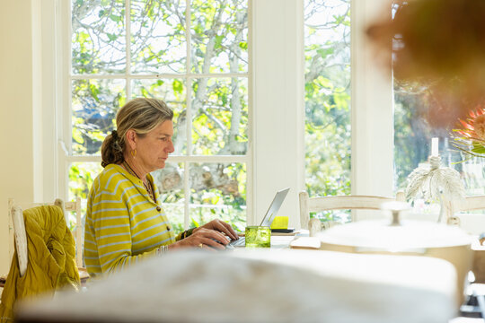adult woman sitting at table in light filled room using mobile phone and typing on laptop on table