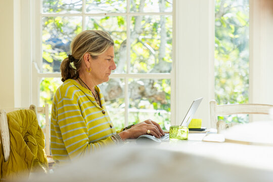 adult woman sitting at table in light filled room typing and using mobile phone with laptop on table
