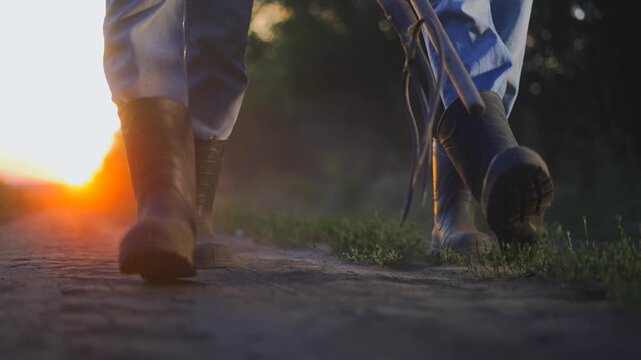 Farmer boots pitchforks road. A man walks along a dirt path. The man wears sturdy boots as the sun sets in the background, radiating soft light.