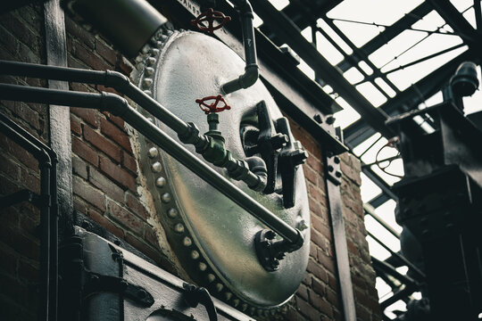 Industrial boiler machinery with red valves mounted on a rustic brick wall in a factory setting