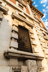 Ornate stone facade of a historic palazzo with a barred window and carved rustication seen from below against a blue sky with clouds, vertical low angle view of classic European architecture in golde © daily_creativity