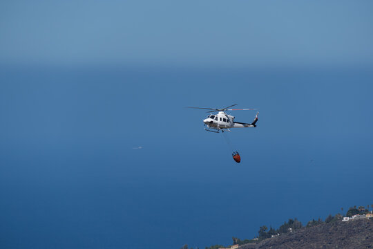 Firefighting helicopter carrying water bucket against ocean background during emergency response, La Palma