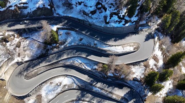 serpentine switchback road snakes down mountainside stacked hairpin loops rocky slope patchy snow sparse pine forest maloja pass straight down aerial drone footage graubunden switzerland