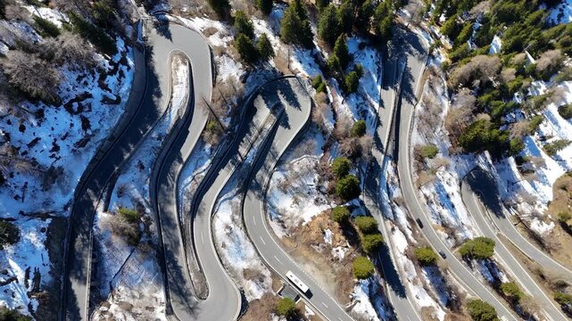 hairpin loops stack down forested mountainside bus truck cars navigate switchback bends patchy snow maloja pass overhead drone footage graubunden switzerland val bregaglia