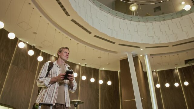 Low angle shot of young adult man standing in modern hotel lobby unfolding foldable smartphone. Perfect for smart travel apps, mobile technology, tourism, and hospitality marketing campaigns
