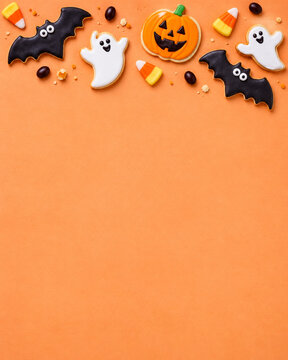Halloween decorative top border with pumpkin, ghost and bat cookies, candy corn and festive treats on an orange background with generous copy space