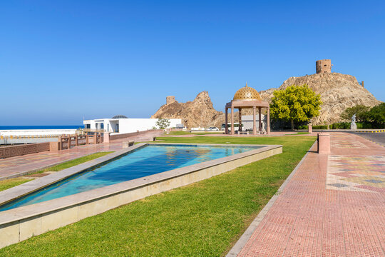 A small park and water feature with the historic watchtowers behind along the corniche waterfront in Al Riyam Park, Muscat, Oman.