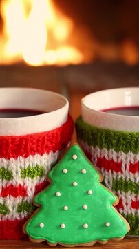 Mugs in knitted woolen covers with a Christmas ornament are placed in front of the fireplace. Xmas decorations on a wooden table in front of a flame burning in the fireplace. Vertical video