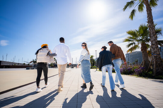 Bright outdoor scene of multicultural friends strolling away together across a modern promenade with palm trees, blue sky