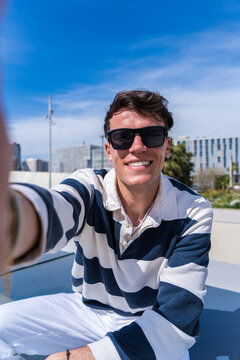 Vertical photo of a young man in sunglasses taking a selfie outdoors while sitting in a bright modern urban setting with blue sky and palm trees.