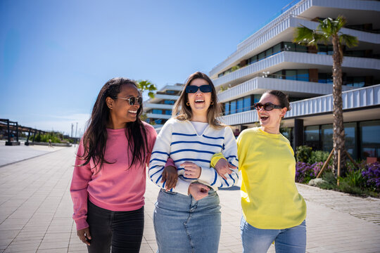 Female friends enjoy a cheerful walk together on a sunny day in a clean residential promenade with palm trees and contemporary buildings.