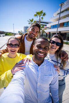 Vertical. Four cheerful friends gather closely for a selfie in a bright residential promenade, enjoying friendship and a sunny day together outdoors.