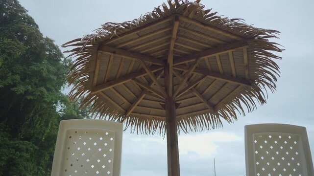 Natural thatch covers the wooden umbrella frame. White lattice panels stand beside the shelter. Lush green trees frame the overcast sky backdrop. This setup invites outdoor relaxation