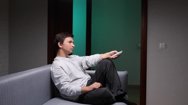 Young man relaxing on a gray couch in a dim living room, holding a remote and watching tv, enjoying quiet evening downtime and simple entertainment at home