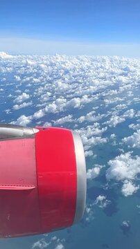 Airplane wing with red engine above fluffy white clouds and blue ocean, clear sky visible in the background. 4K vertical footage