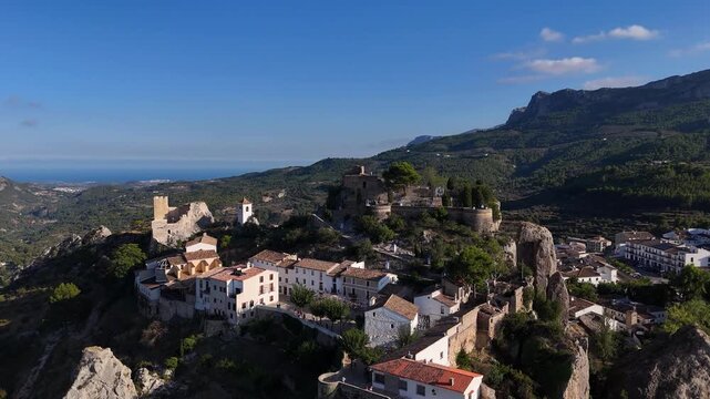 Aerial view of the medieval castle village of Guadalest, Alicante, Spain, perched on a rocky crag, with terracotta roofs, stone walls, the Penon de Ifach and the sea on the horizon