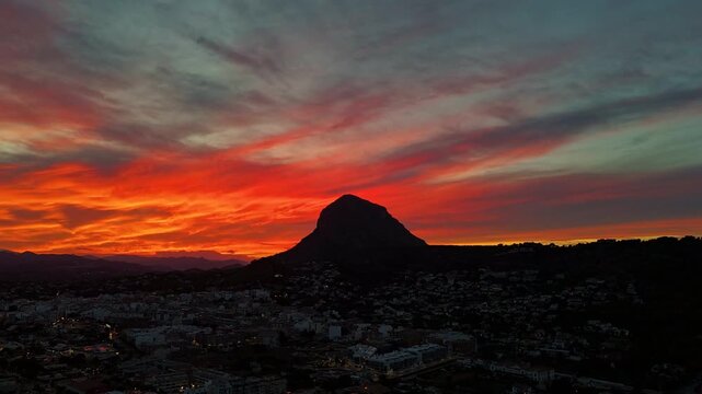 Hyperlapse drone footage of Montgo mountain silhouetted against a fiery sunset over Javea, camera pushing forward and pulling back across town lights, Alicante, Spain