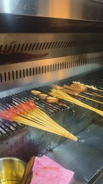 A chef grills skewers of marinated meat, mushrooms, and other ingredients over charcoal on a commercial barbecue grill, with smoke rising from the fire at vertical composition
