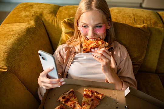 Young woman eating pizza on sofa while using smartphone at home