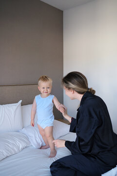Happy toddler standing on a bed while interacting with his mother in a cozy bedroom. Candid family lifestyle moment showing motherhood, childhood, play and parent-child bonding.