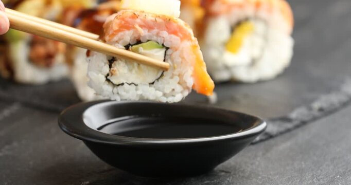 Woman dipping tasty sushi roll into soy sauce at black table, closeup