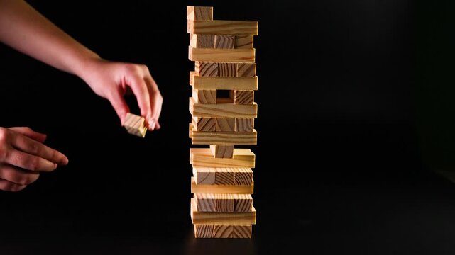 Players taking turns removing wooden blocks from unstable tower in strategy board game against black background.