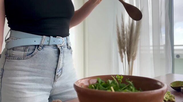 Mid adult millennial woman preparing a healthy green salad at home while drinking wine, dancing and singing, enjoying a relaxed and playful moment during everyday cooking.