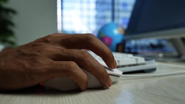 Hand Using Computer Mouse at Home Office Desk, Close-up of a person's hand using a computer mouse beside a keyboard and monitor in a home office workspace.