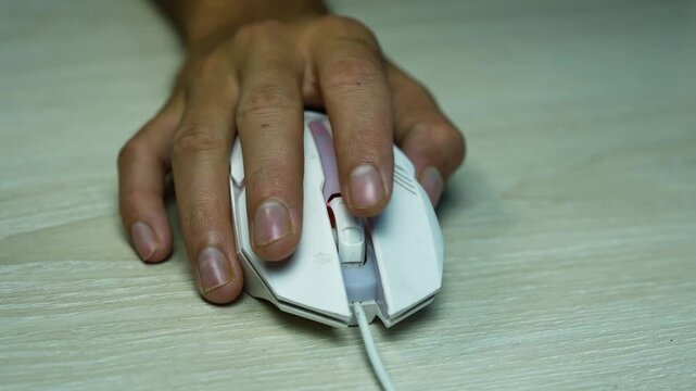 Hand using a wired computer mouse on desk, Close-up of a hand using a white wired computer mouse on a light wooden desk, showing everyday office work and technology use.