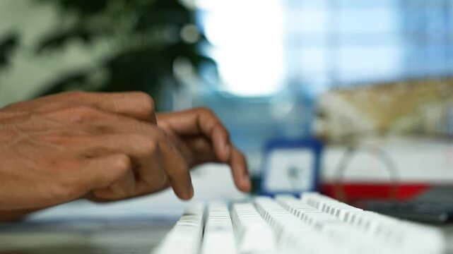 Hands typing on a computer keyboard at office desk, Close-up of hands typing on a white computer keyboard at a desk, with a blurred office background and shallow focus.