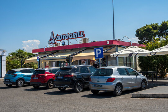 Milazzo - ME - IT - Sep 09, 2026 : Autogrill roadside restaurant with parked cars, outdoor umbrellas, awnings and a blue parking sign under a clear sky. Trees and palms border the paved parking area