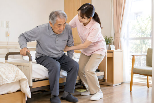 Female caregiver assisting an elderly Japanese man to stand from bed