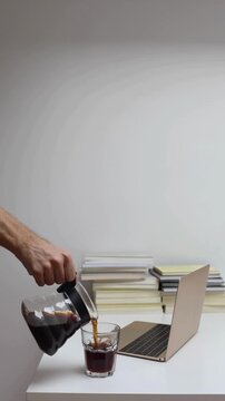 Hands Pouring Coffee into Cup Beside Laptop and Stack of Books on Desk